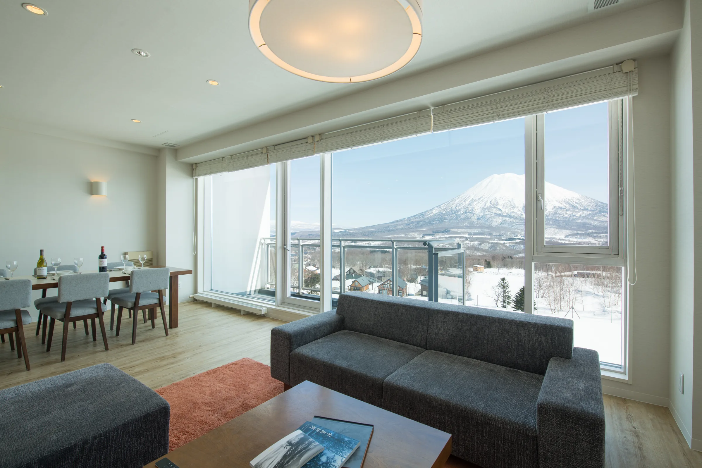 Open-plan living space with dark sofa, dining table and floor-to-ceiling windows framing snow-capped mountain view.